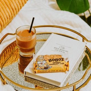 A tray with a glass of chocolate and a book, alongside a package of Bobo’s Banana Chocolate Chip Oat Bars