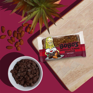 A bowl of chocolate chips, a package of oat bars, and a plant with nuts. Close-up of a bag of brownies and a peanut. Group of almonds on a purple surface.