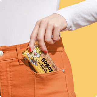 Image of a person wearing orange pants holding a Bobo's Banana Chocolate Chip Oat Bar on a yellow background